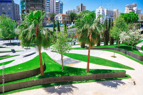Obraz na płótnie Modern urban park in Nicosia city, with green lawns, palm trees, and other vegetation, set against a backdrop of various buildings. Park features curving pathways and concrete structures w salonie