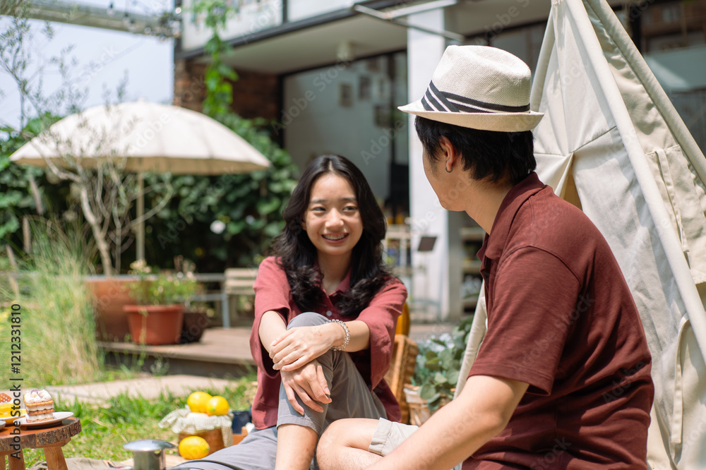 An Asian couple enjoying a joyful picnic outdoors. The man playfully offers a cupcake to the smiling woman. They sit on a blanket near a tipi, surrounded by snacks and a lush garden backdrop An Asian couple enjoying a joyful picnic outdoors. The man playfully offers a cupcake to the smiling woman. They sit on a blanket near a tipi, surrounded by snacks and a lush garden backdrop