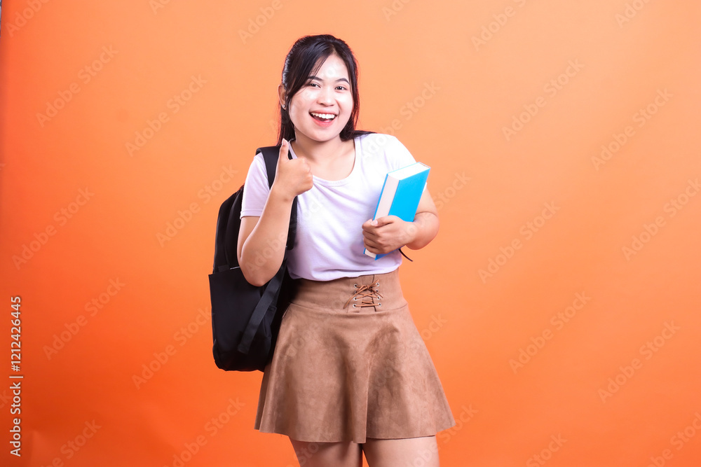 Fototapeta premium Young woman with book and backpack gives thumbs up gesture on an orange background