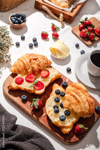 Delicious breakfast spread featuring buttery croissants topped with fresh strawberries and blueberries, served on a wooden board with butter, coffee, and fruit, creating a cozy