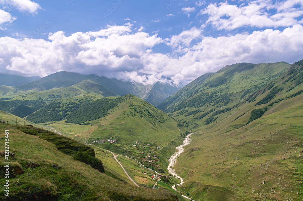 Obraz premium Panorama in the mountains with a view of the village and the mountain river. Water flows down from the peaks of the mountains, forming a river. There are white clouds over the mountains.