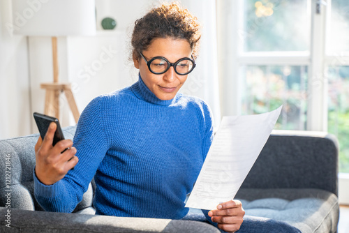 Black woman reading document and holding smartphone at home photo