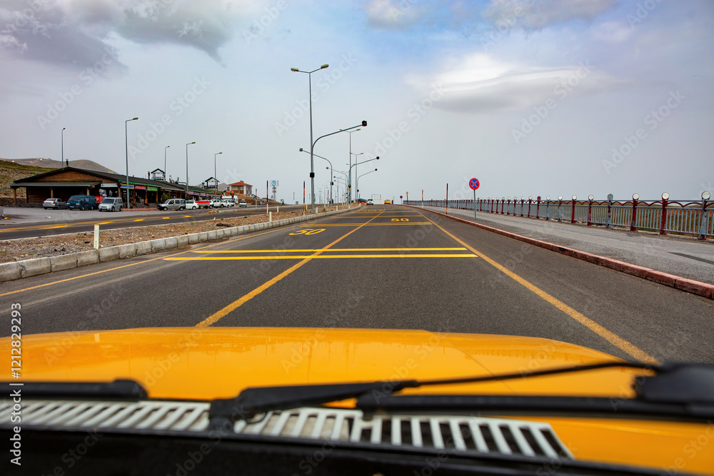 Journey down a coastal road with yellow car and distant mountains under a cloudy sky