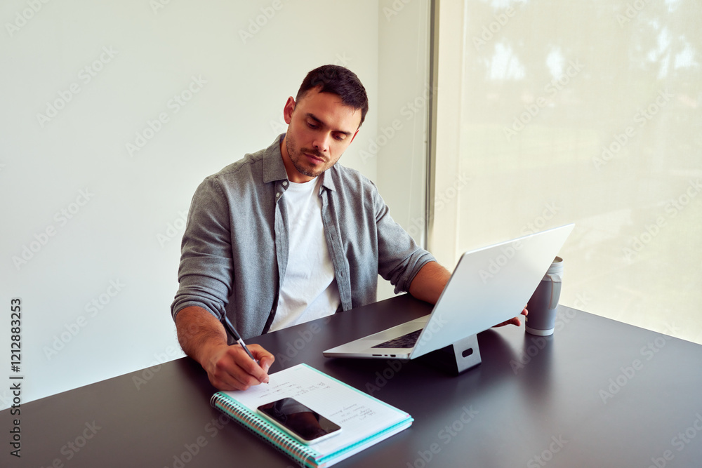 Young businessman taking notes in notebook while working on laptop in bright modern office
