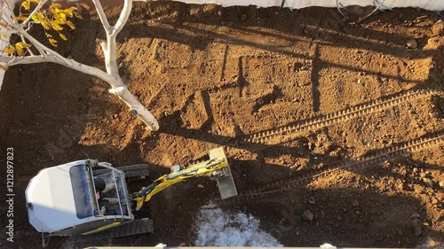 An excavator operates in a yard, leveling soil during ongoing construction, with visible tire tracks and a tree nearby.
