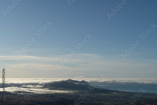 A wall of mist rolls in from the sea towards mountains wrapped in a cloud inversion under a clear blue sky. photo