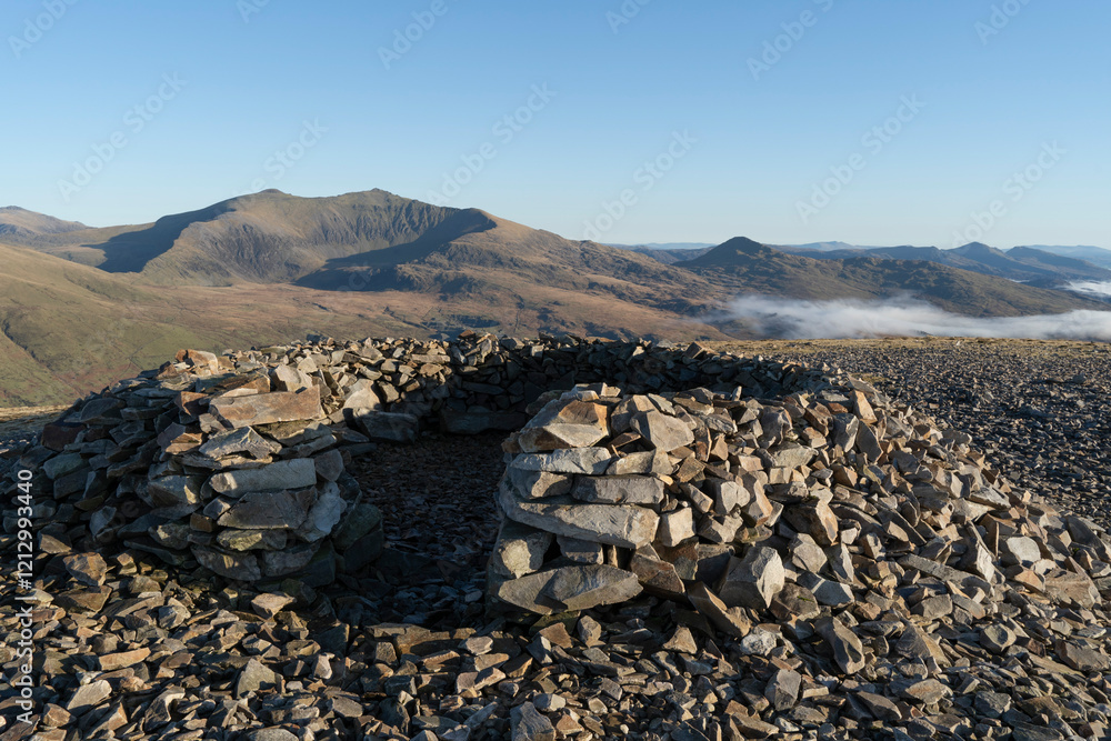 Φωτογραφία Circular stone cairn shelter with a vista of mount Snowdon, Wales, UK, in the distance under a clear blue sky