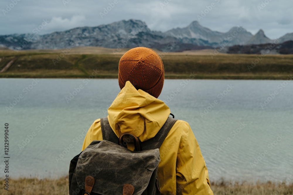 A young traveler woman wearing yellow raincoat and retro style backpack, hiking in mountains and enjoying views of nature. Rear view. Vrazje jezero - Montenegro country, Durmitor national park. A young traveler woman wearing yellow raincoat and retro style backpack, hiking in mountains and enjoying views of nature. Rear view. Vrazje jezero - Montenegro country, Durmitor national park.
