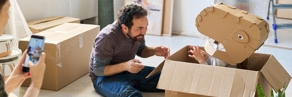 Header of man sitting on the floor and looking at little daughter in cardboard dinosaur mask in front of woman taking picture of them Header of man sitting on the floor and looking at little daughter in cardboard dinosaur mask in front of woman taking picture of them
