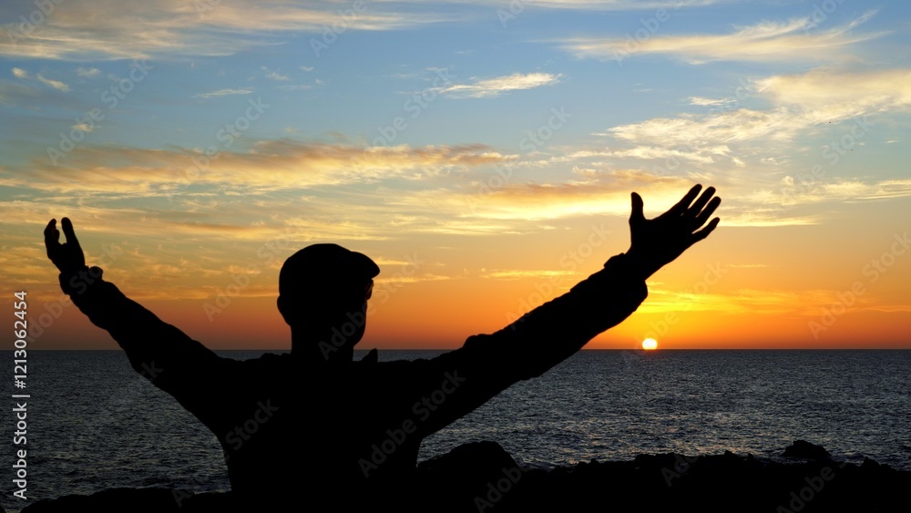 Fototapeta premium Silhouette of a young traveler raising arms in front of a beautiful sunset over the ocean in Canary Islands, celebrating freedom and enjoying the moment of solitude