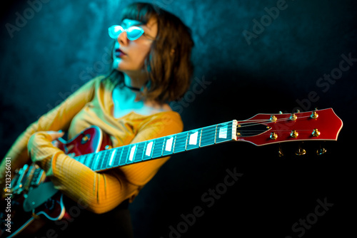 Fashion young hipster woman with curly hair with red guitar in neon lights. Rock musician posing with electrical guitar.