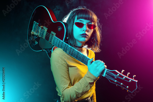 Trendy young hipster woman with curly hair with red guitar in neon lights. Rock musician posing with electrical guitar.