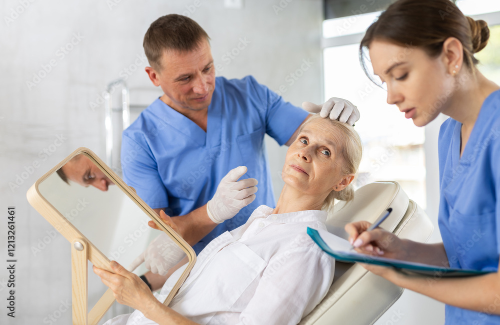 Obraz premium Interested elderly woman sitting on examination couch and looking at mirror while professional cosmetologist with female assistant planning and explaining future rejuvenating facial procedure