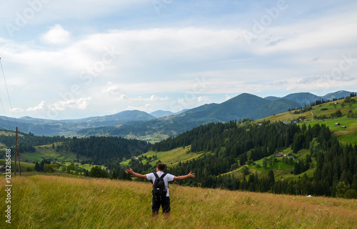 Serene landscape featuring a man with arms outstretched in a lush mountain valley, surrounded by vibrant green forests and rolling hills under a picturesque clear sky. Carpathians, Ukraine