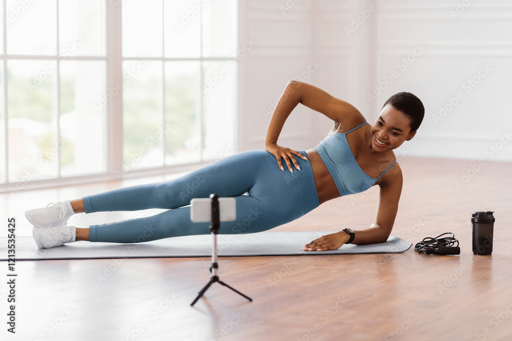 Obraz premium Online Workout. Smiling African Lady Standing In Side Plank Position On Yoga Mat Near Window, Looking At Mobile Phone On Tripod, Watching Tutorial Or Recording Video For Blog, Having Chat With Coach