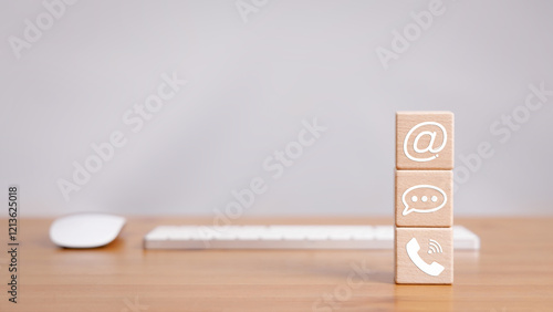 Cube wood block stacked on wooden table with phone, message bubble, and email sign icon with computer keyboard and mouse in contact us concept.