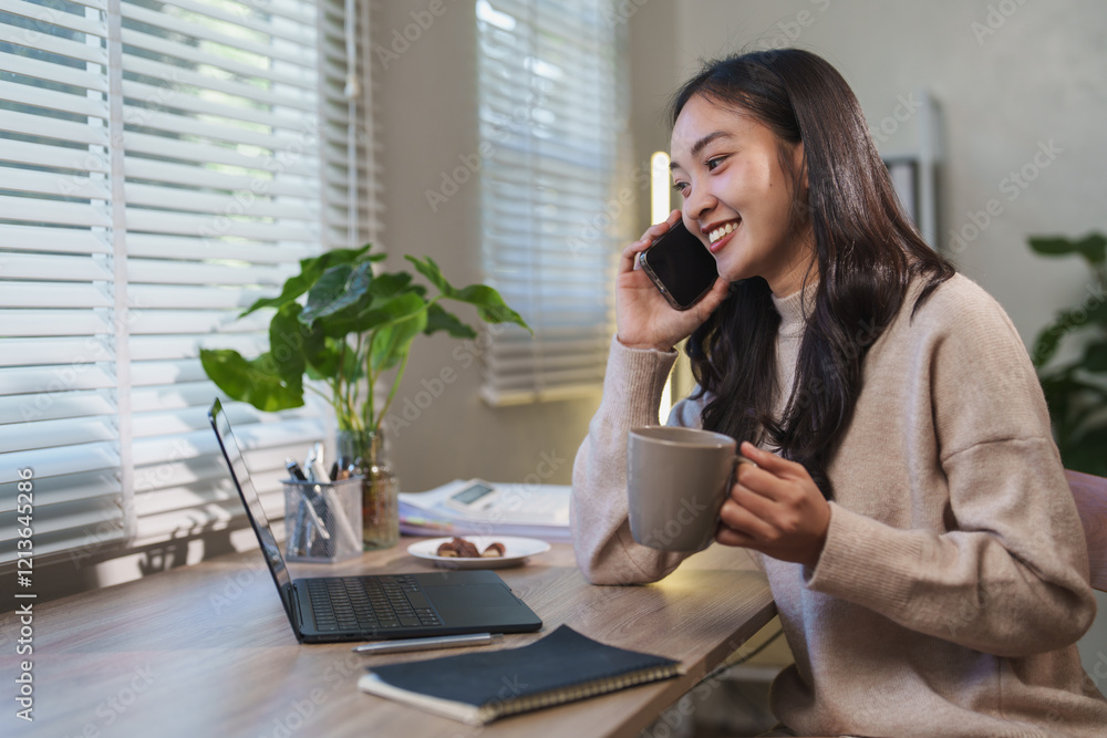 Young asian woman working from home, chatting on the phone and sipping coffee while seated at her desk with a laptop and notebook, enjoying a cozy and relaxed morning Young asian woman working from home, chatting on the phone and sipping coffee while seated at her desk with a laptop and notebook, enjoying a cozy and relaxed morning