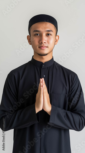 A portrait of a young asian Muslim man wearing black traditional Islamic attire, making an apologetic gesture with his hands, isolated white background photo