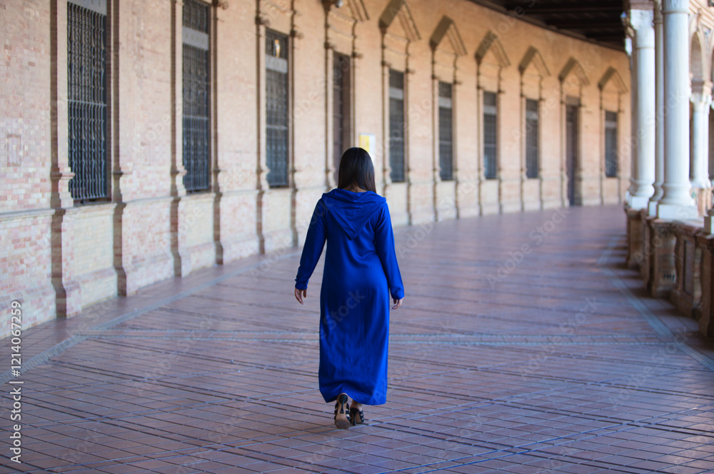 Naklejka premium moroccan brunette woman in blue abaya, typical arabian costume, walking between arches and columns in the spain square in seville. Photo taken from behind. Travel and holiday concept.