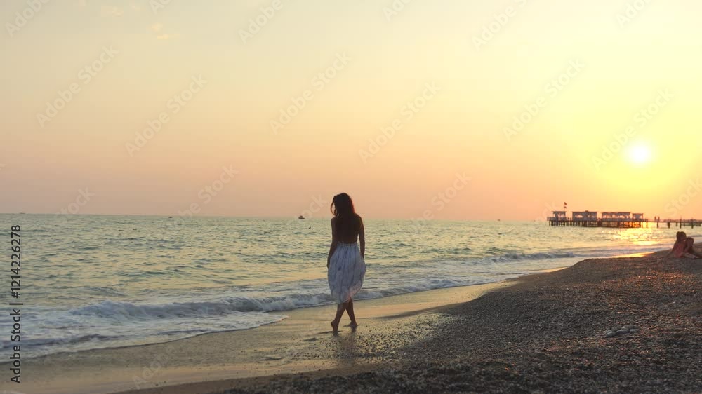 A young woman walks along a quiet beach at sunset, enjoying the peaceful sea breeze and golden light reflecting on the water.