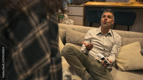 Couple argues. A gray-haired man sits on the couch with a joystick, while his girlfriend throws dirty clothes at him, frustrated that he hasn't cleaned up. She is angrily scolding him photo