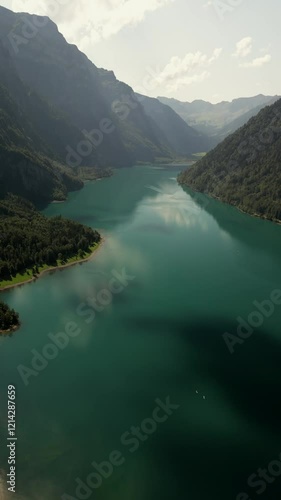Vertical drone shot of beautiful glacial emerald-green lake surrounded by lush forests, natural lake in summer afternoon under clear blue sky