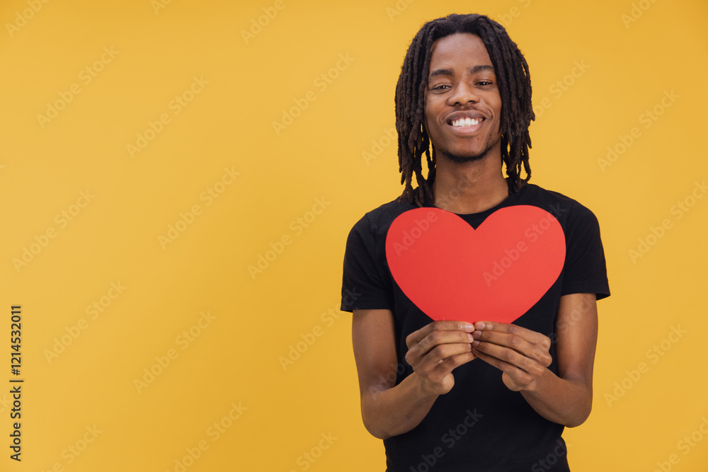 Young man holding a big red heart, smiling on yellow background Young man holding a big red heart, smiling on yellow background