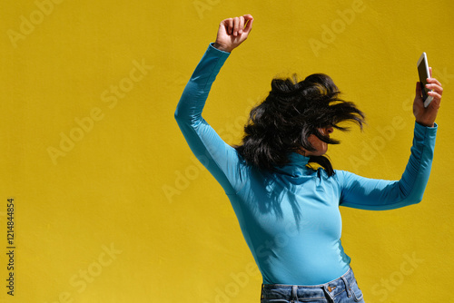 Young stylish woman dancing and holding smartphone against yellow wall photo