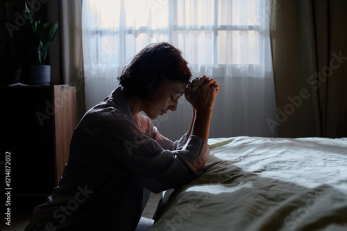 Картината върху платно Side view of woman in pajamas kneeling near made bed holding her hands together