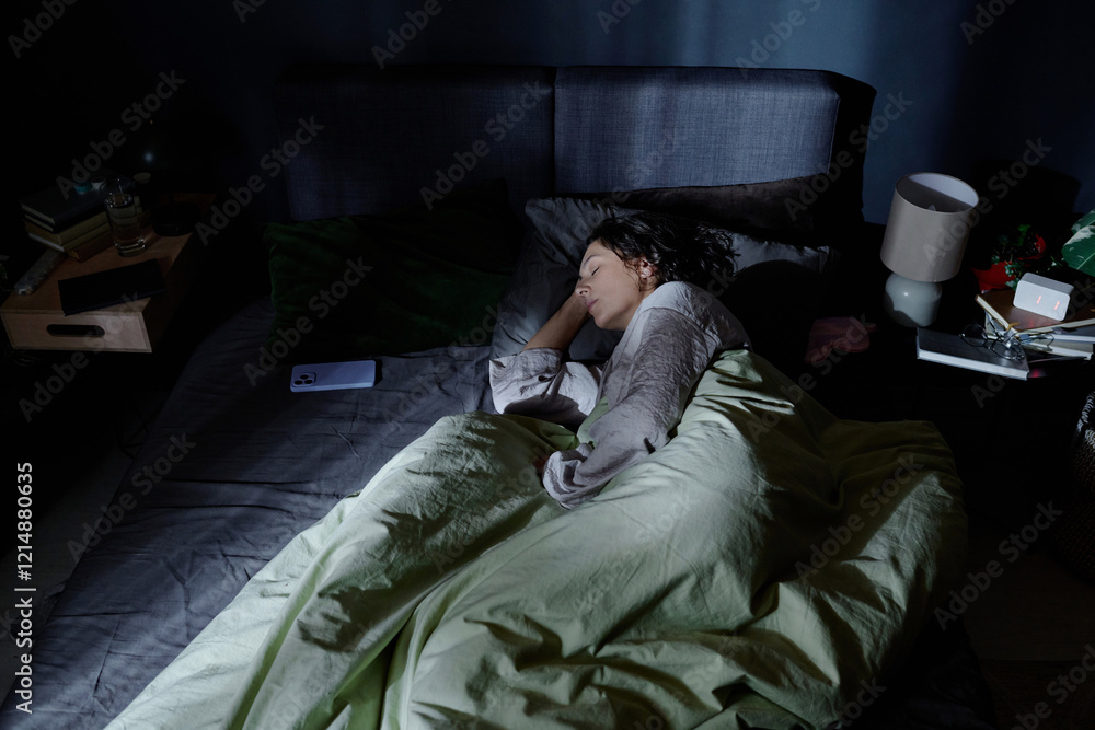 Fototapeta premium High angle shot of sleeping woman with olive blanket enjoying rest time in comfortable bed while moonlight illuminating her bedroom