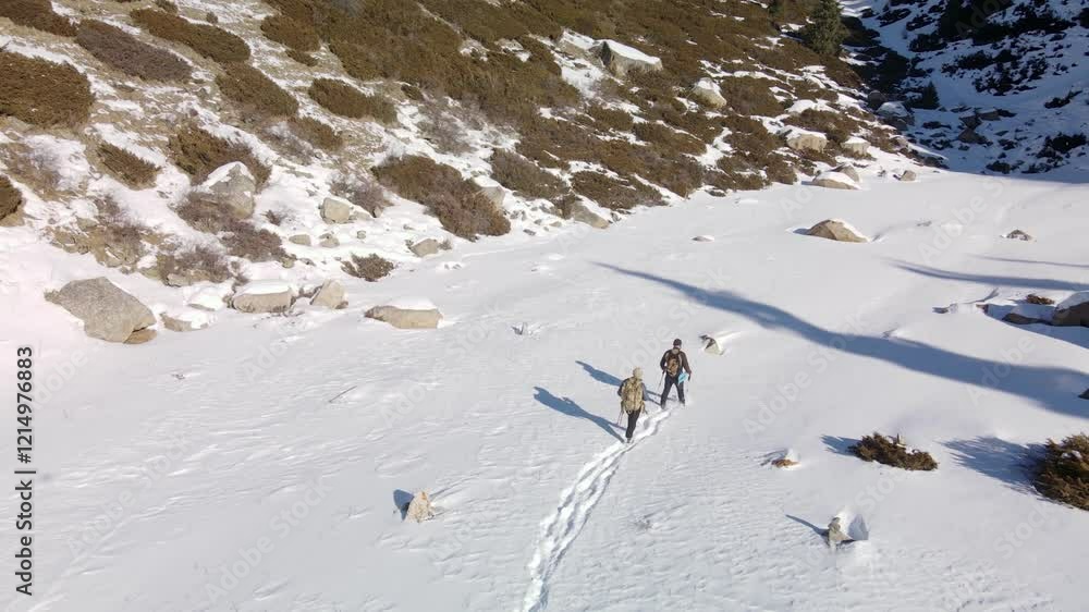 Two tourists with backpacks walk along a snowy valley in the mountains, trampling a path. Aerial view of two tourists with backpacks in the mountains in winter. Two tourists with backpacks walk along a snowy valley in the mountains, trampling a path. Aerial view of two tourists with backpacks in the mountains in winter.