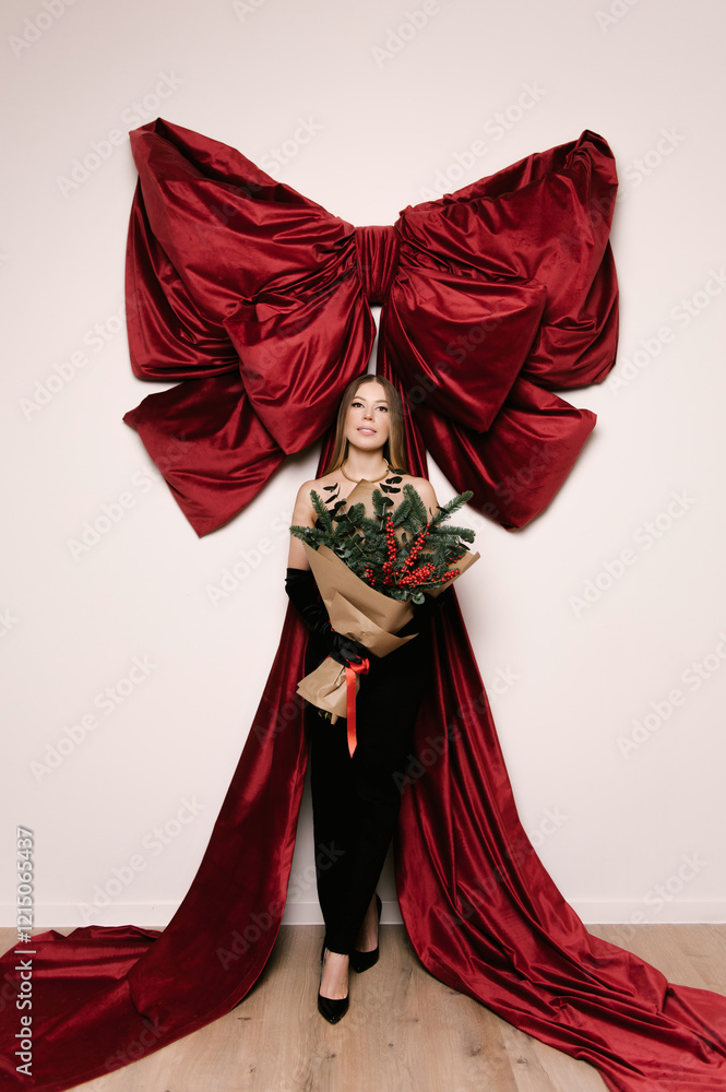 Portrait of a woman in a studio with professional lighting in a black elegant dress with a Christmas tree bouquet against the background of a large red bow
