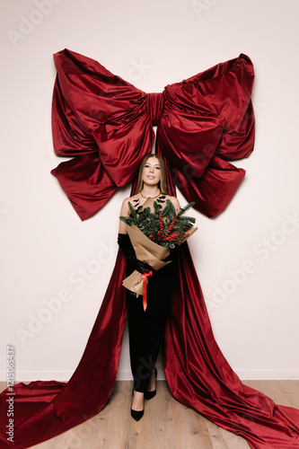 Obraz na płótnie Portrait of a woman in a studio with professional lighting in a black elegant dress with a Christmas tree bouquet against the background of a large red bow w salonie