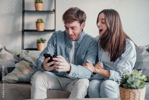 Excited millennial husband and wife relax on couch in kitchen feel euphoric win lottery online on cellphone, overjoyed young couple triumph celebrate read good unexpected news on smartphone gadget.