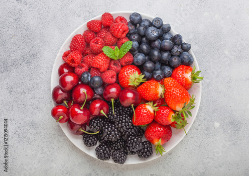 Fresh organic summer berries mix in white plate on light kitchen table background. Raspberries, strawberries, blueberries, blackberries and cherries.