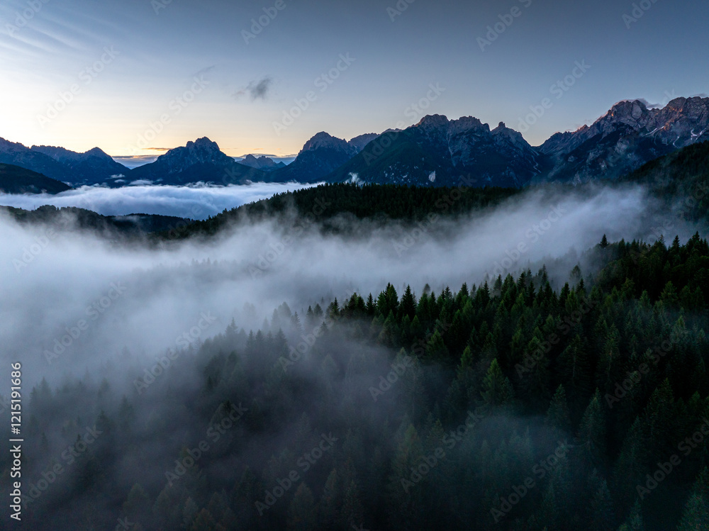 A mist-filled valley in the Dolomites at sunrise, with sunlight highlighting the peaks and dense alpine forest below. Taken near Danta village, Italy. Aerial photo with Dolomites. Hike in Italy.