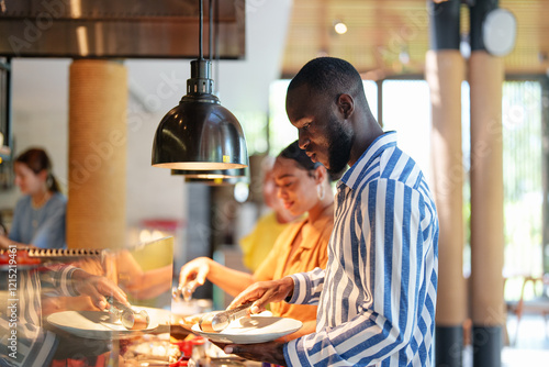 Obraz na plátně A multiracial female and a Black male, both young adults, are seen choosing food in a modern self-service restaurant