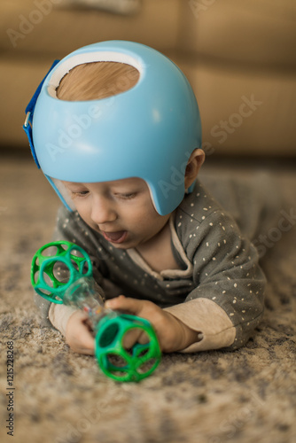 a baby boy wearing a blue Helmet for molding therapy, for cranial orthosis for plagiocephaly while playing with a toy on the carpet