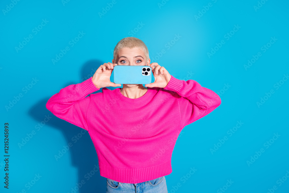 Young woman with short blonde hair holding a smartphone in front of her face, wearing a vibrant pink sweater against a blue background Young woman with short blonde hair holding a smartphone in front of her face, wearing a vibrant pink sweater against a blue background