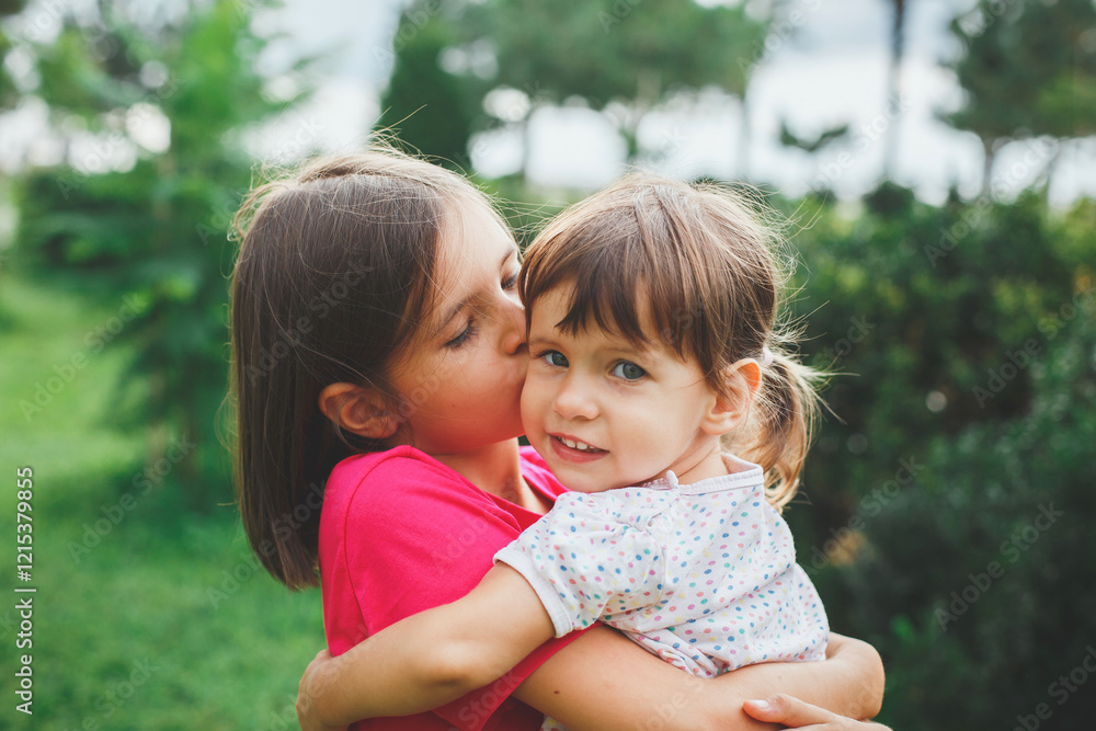 Older sister lovingly kisses her younger sister on the cheek while hugging her outdoors in a lush green park, symbolizing family bonds, childhood, and affection, summer portrait Older sister lovingly kisses her younger sister on the cheek while hugging her outdoors in a lush green park, symbolizing family bonds, childhood, and affection, summer portrait