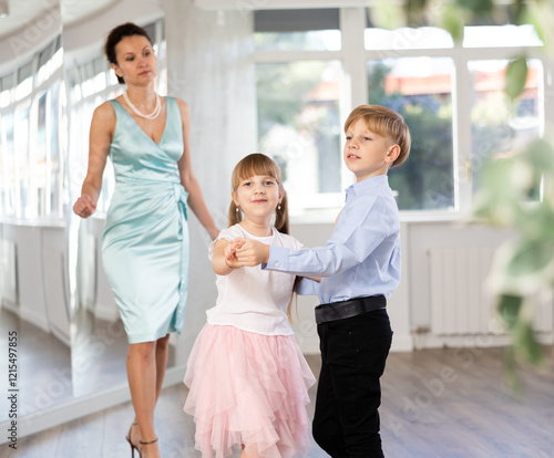 Small passionate ballroom enthusiasts, tween girl and boy practicing graceful waltz routine in well-lit dance hall under female mentor watchful eye.. photo