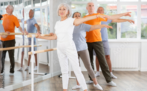 Pensioners of different ages exercise near ballet barre at group training session in the two position photo