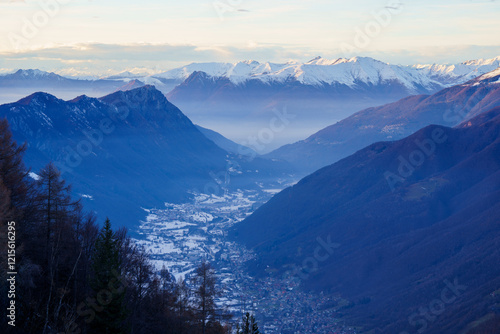 Snowy village is nestled in mountain valley at dusk, surrounded by towering peaks and mist, creating peaceful and remote alpine scene near Piani di Bobbio, Italy.