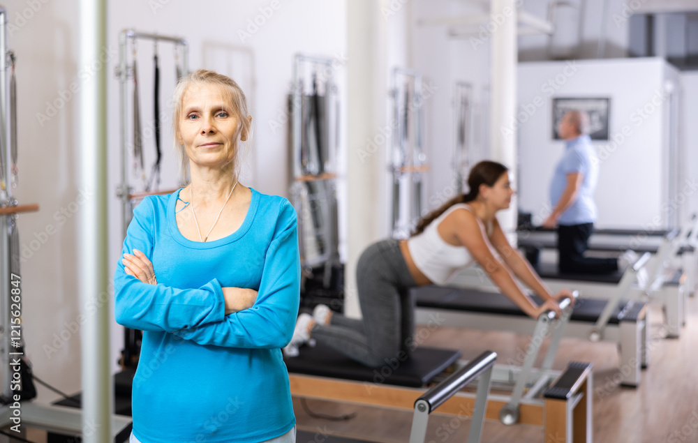 Obraz premium Portrait of smiling mature woman posing against women exercising on reformer bed machine in Pilates studio