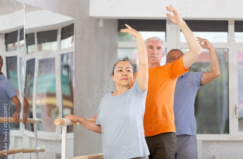 Elderly women and men practice at ballet standing in fourth position at barre in bright choreography studio photo