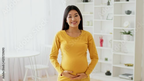 A joyful Asian expectant mother stands in a well-lit living room, cradling her baby bump. She wears a bright yellow top and exudes happiness surrounded by a modern, cozy interior.