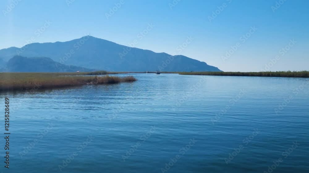 Moving shot from a boat over the Azmak River Delta in Dalyan, Turkey, showcasing the crystal-clear blue waters, reeds along the shore, and the majestic mountains in the background. Moving shot from a boat over the Azmak River Delta in Dalyan, Turkey, showcasing the crystal-clear blue waters, reeds along the shore, and the majestic mountains in the background.