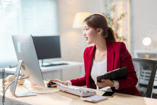 A woman in a red suit is sitting at a desk with a computer monitor photo