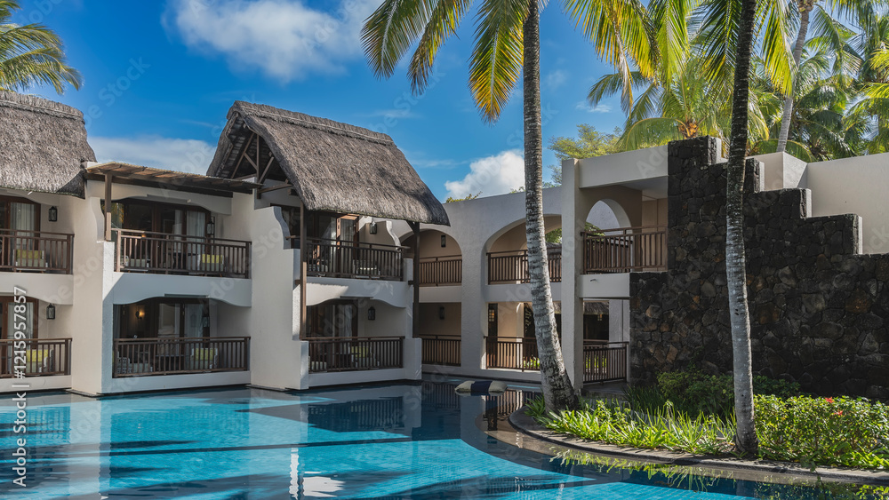 Fototapeta premium Swimming pool at the resort. Cottages with thatched roofs and terraces around the perimeter. Palm trees, green grass in a flower bed. Shadows on clear aquamarine water. Blue sky, clouds. Mauritius.