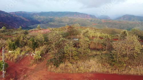 UHD Kauai, Hawaii: Aerial View of White RV on Scenic Dirt Road Amidst Majestic Mountains.Immerse in the Richness of Verdant Green and Earthy Brown Vegetation, Accented by Patches of Striking Red Soil. photo